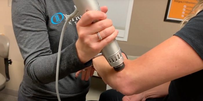 A female physical therapist applies shockwave therapy to arm of female patient near the elbow, using a handheld Radial Pulse Therapy tool to alleviate pain due to plantar fasciitis.